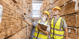 Two workers wearing safety attire look at stacks of lumber in a warehouse