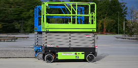 Green and blue electric scissor lifts outside in a parking lot in front of trees