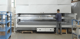 A worker using a large vertical lift module on a warehouse floor with shelving racks in the foreground